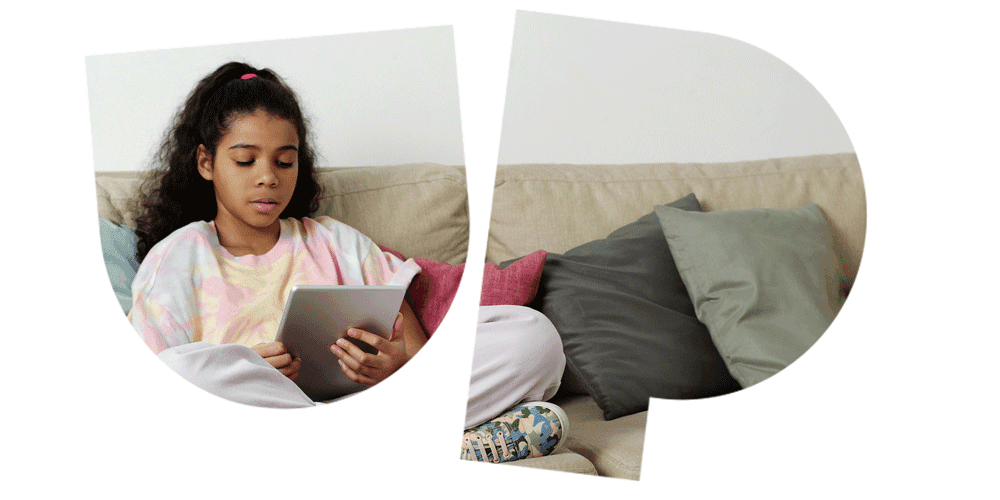Girl with curly hair reading on a tablet while sitting on a beige sofa with colorful cushions.