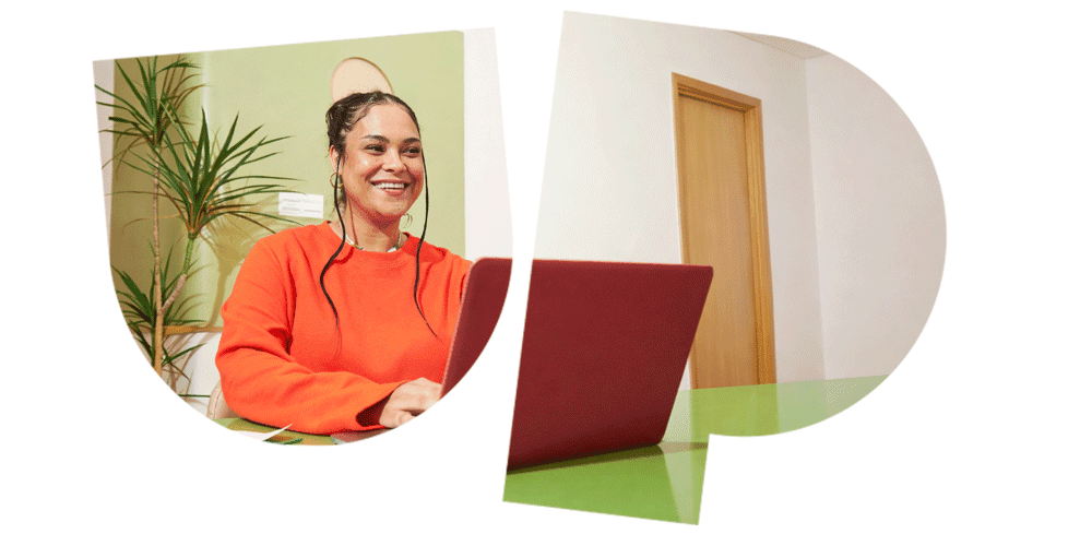 Woman in orange sweater smiling while using a red laptop, sitting at a green desk with a plant and wooden door in the background.
