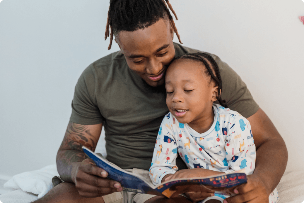 Father and child reading a book together, smiling and bonding. The child wears colorful pajamas, seated on the father's lap.
