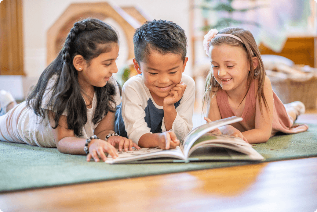 Three children lying on a green carpet, smiling and looking at a book together in a cozy room.