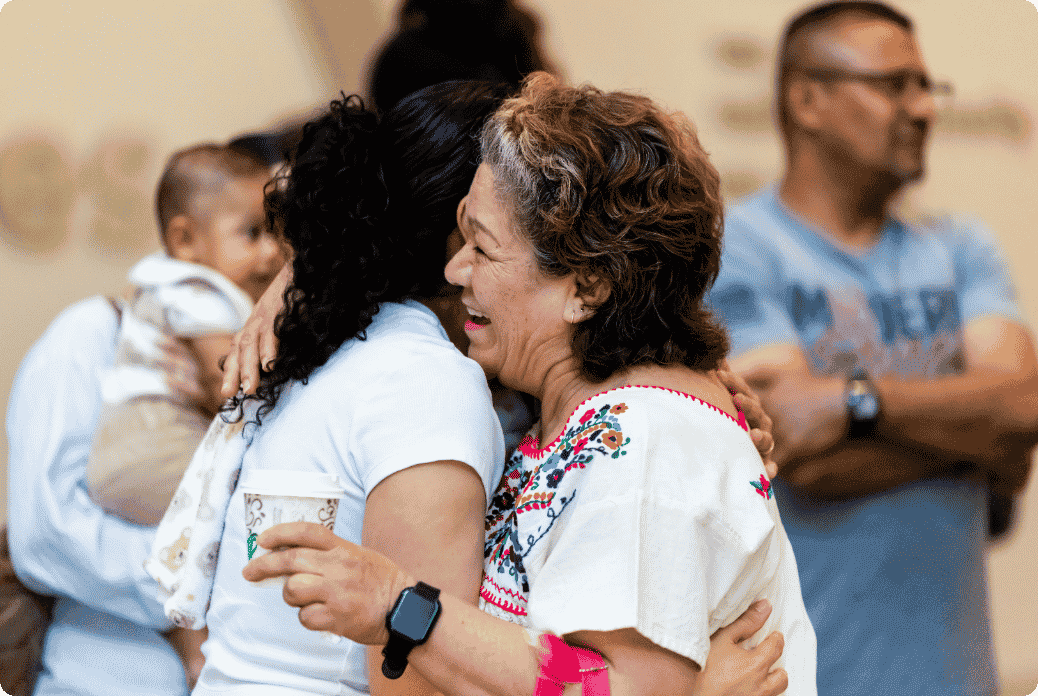 Elderly woman in embroidered blouse joyfully hugs a person while holding a coffee cup, with people chatting in the background.