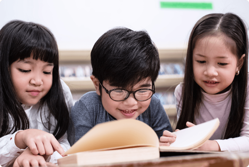 Three children happily read a book together, with one child pointing at the pages while smiling.