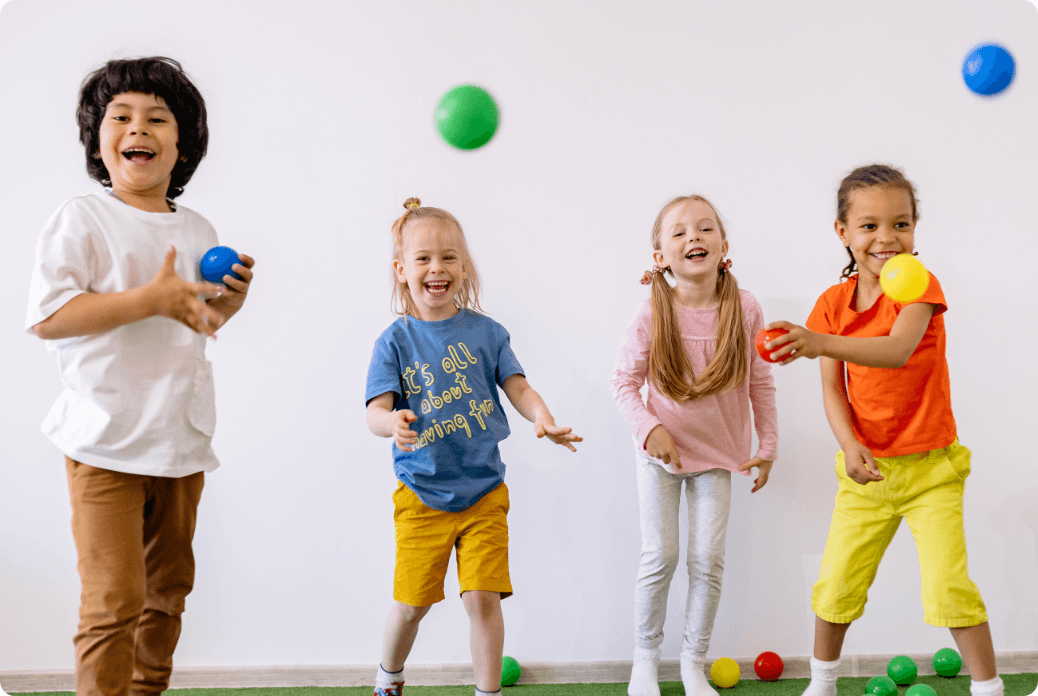 Four children joyfully play with colorful balls indoors, smiling and wearing casual, vibrant clothing.