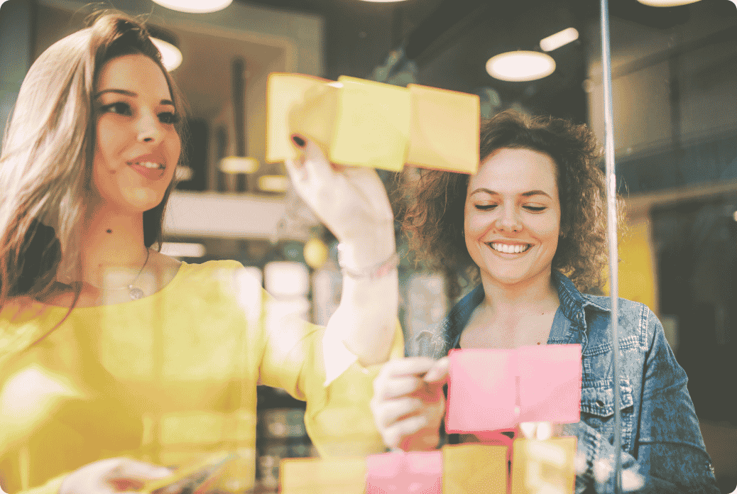Two women smiling and sticking colorful post-it notes on a glass wall in a bright, modern office setting.