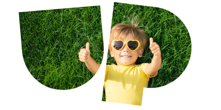 Child lying on grass, wearing a yellow shirt and heart-shaped sunglasses, giving thumbs up.
