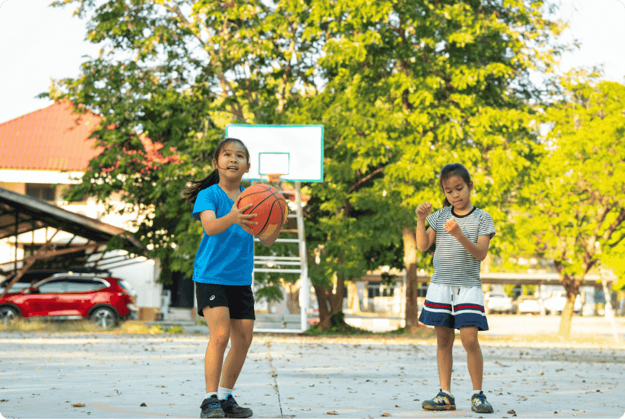 Two young girls play basketball outdoors on a sunny day, one holding a ball and smiling, with trees and a hoop in the background.