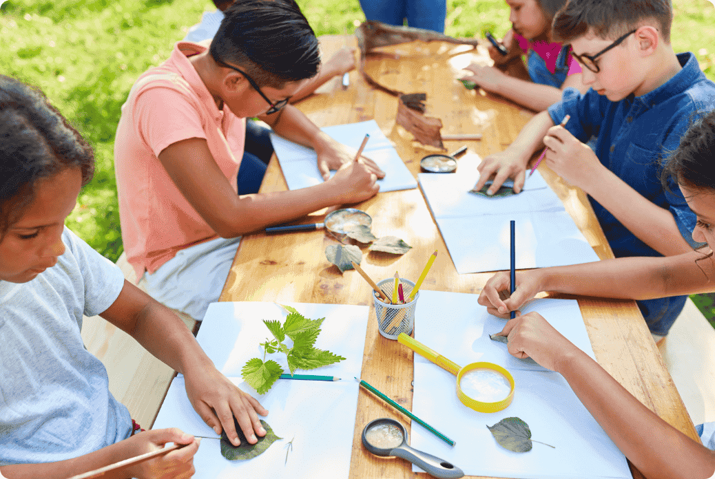 Children sit at a wooden table outdoors, studying leaves with pencils and magnifying glasses on paper, engaging in a nature-based activity.