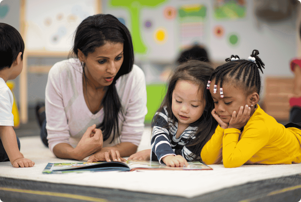 A teacher and two children are lying on a carpet, reading a book together in a classroom.