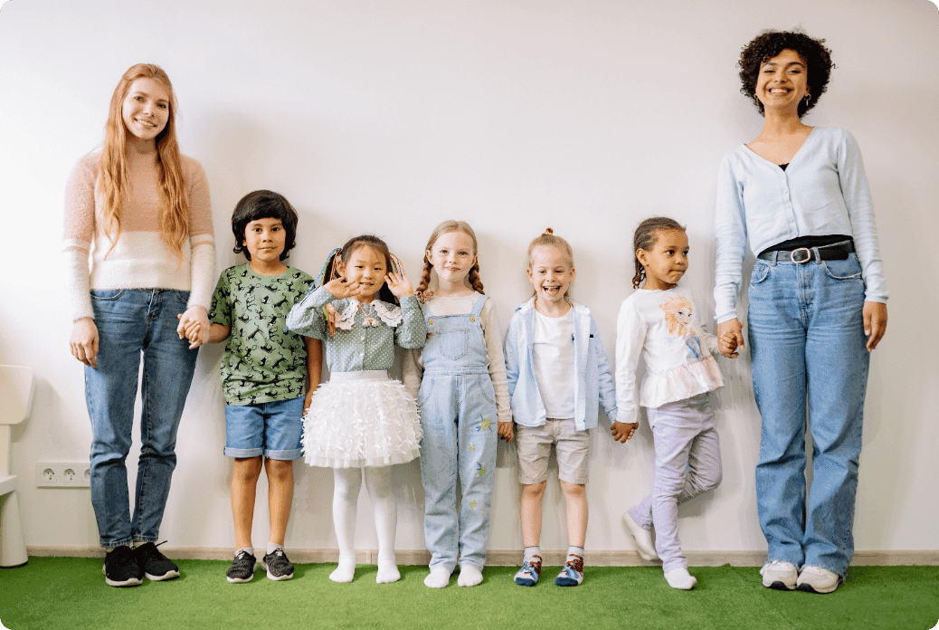 Two women and five children stand against a white wall, smiling and holding hands, on a green carpet.