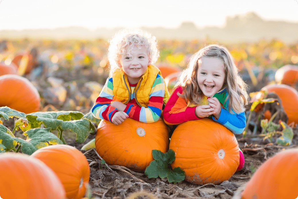Two smiling children in colorful striped sweaters leaning on large pumpkins in a sunlit pumpkin patch.